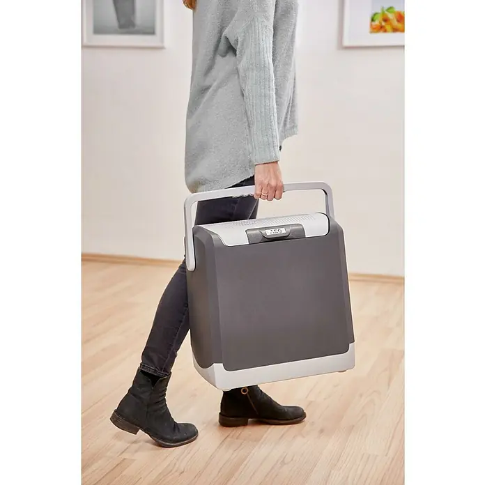 AEG Nevera de camping KK 20 - Capacidad volumétrica: 20 l, Gris - Mujer cargando una nevera portátil. La nevera es gris con un asa y detalles en blanco y tiene una pantalla digital.