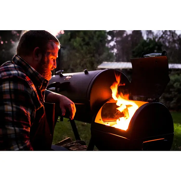 Char-Broil Smoker Oklahoma Joe - Grillfläche (B x T): 88 x 44 cm - Mann am Smoker mit offenem Feuerraum. Der Smoker ist aus Metall und hat einen Garraum und einen Feuerraum für das Holz.