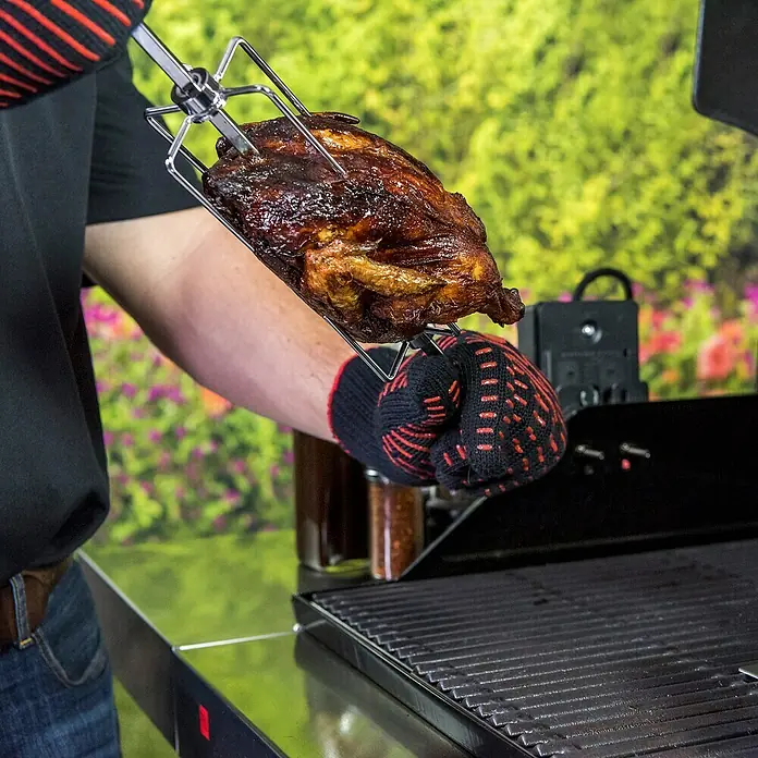 Char-Broil Drehspieß - Länge: 70 cm - Ein gegrilltes Hähnchen am Spieß wird mit hitzebeständigen Handschuhen über einem Grill gehalten.