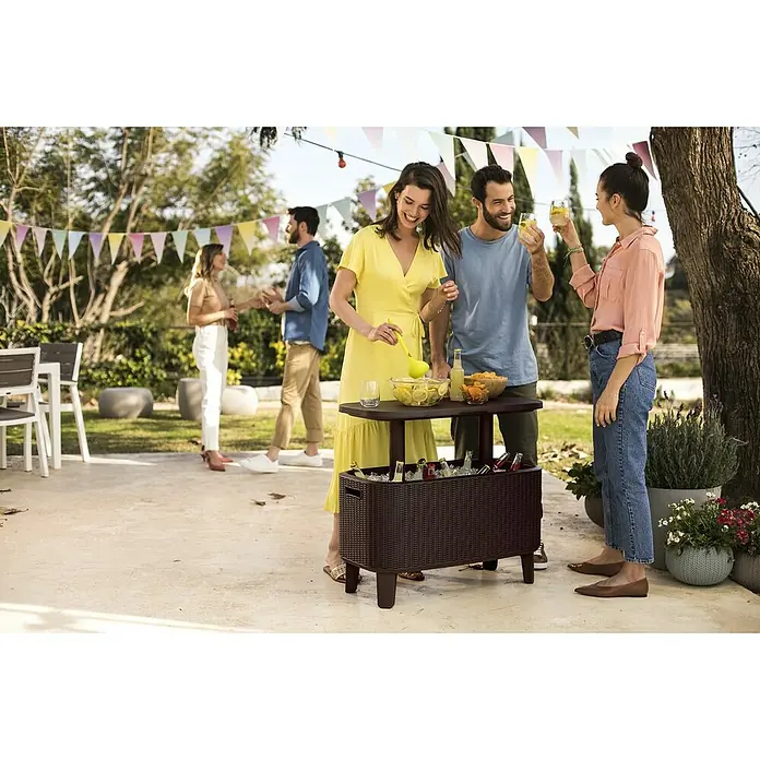 Keter Nevera portátil Bevy Bar - L x An x Al: 83 x 40,5 x 50 cm, Marrón - Un carrito refrigerador marrón con tablero de mesa es el punto focal de una fiesta al aire libre, rodeado de personas celebrando.