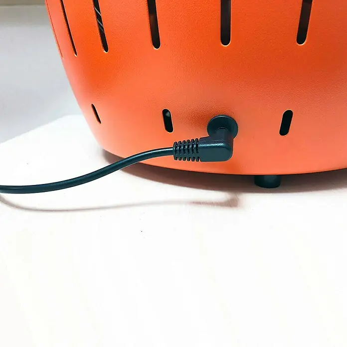 LotusGrill Barbacoa de carbón XL - Diámetro de la superficie de la parrilla: 43 cm, Rojo - Primer plano de una lámpara naranja con un cable de alimentación negro enchufado en la parte posterior. La lámpara tiene un diseño cilíndrico.