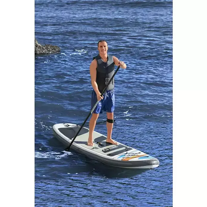 Bestway Hydro Force Paddle surf White Cap - L x An x Al: 305 x 84 x 12 cm, Carga útil: 120 kg, Hinchable, Gris - Un hombre está de pie en una tabla de surf de remo en el agua. La tabla es gris y blanca y el hombre lleva un chaleco salvavidas.