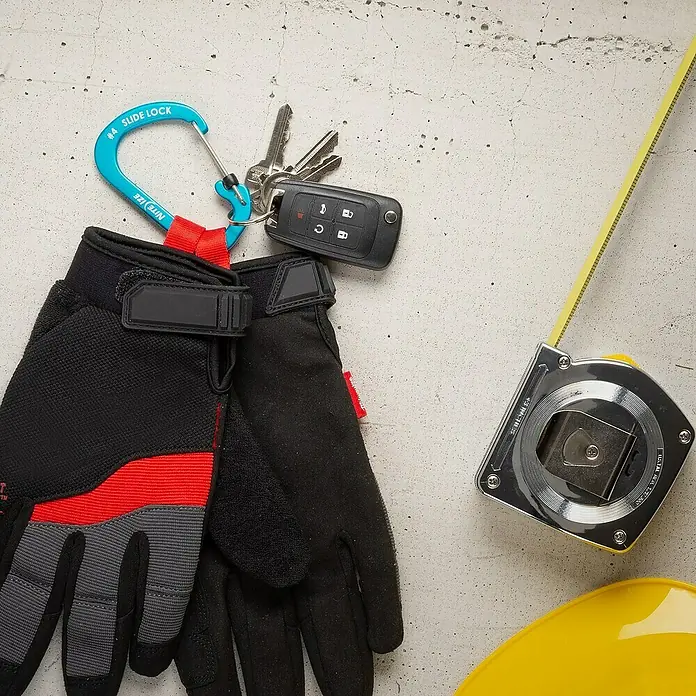 Nite Ize Mosquetón Slide Lock - 81 x 52 mm, Carga soportada: 33 kg - Elementos de trabajo: Guantes de trabajo, un llavero con una llave de coche, una cinta métrica y un casco amarillo sobre hormigón.