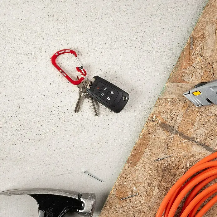 Nite Ize Mosquetón Slide Lock - 58 x 38 mm, Carga soportada: 11 kg - Llavero con mosquetón rojo, llave del coche y llaves de casa sobre un banco de trabajo con martillo, clavos y herramientas.
