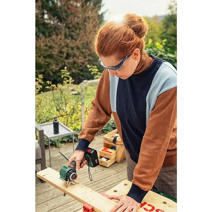 Bosch Power for All 18V Sierra NanoBlade AdvancedCut 18 - Número de revoluciones de marcha en vacío: 7.000 r.p.m., 18 V, 1 batería - Mujer con gafas de seguridad corta madera con una mini sierra circular inalámbrica Bosch conocida por su precisión y fácil manejo.