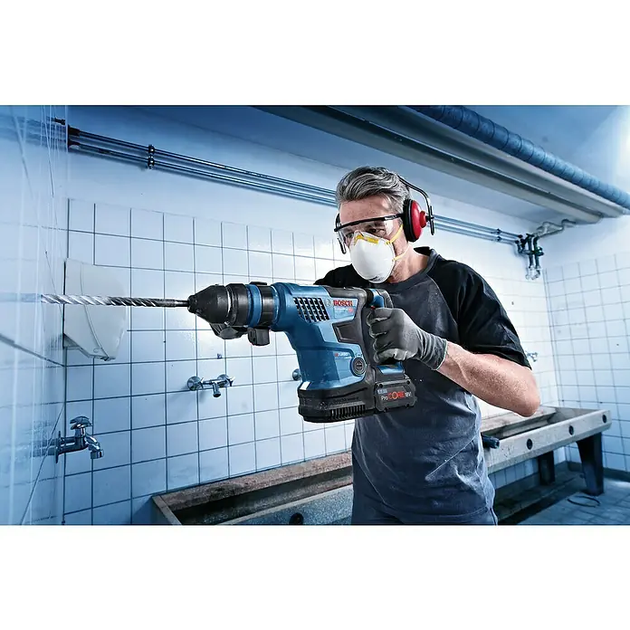 Bosch Professional AMPShare 18V Akku-Kombihammer 18V-34 CF - 18 V, Ohne Akku, 5,8 J - Ein Mann bohrt mit einem blauen Bosch-Bohrhammer in eine Wand. Er trägt Schutzausrüstung und arbeitet in einem Raum mit weißen Fliesen.