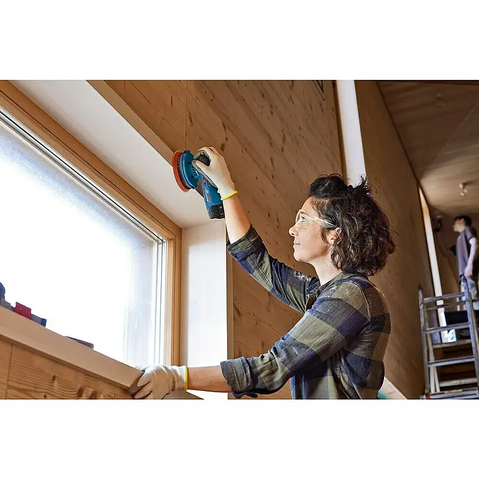Bosch Professional Akku-Exzenterschleifer GEX 12V-125 Solo - 12 V, Ohne Akku, Durchmesser Schleifteller: 125 mm - Frau schleift Holz mit einem blauen Bosch Schleifgerät neben einem Fenster, um es zu veredeln. Im Hintergrund eine Leiter und eine weitere Person.