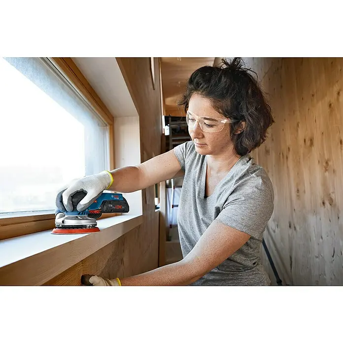 Bosch Professional AMPShare 18V Akku-Exzenterschleifer GEX 18V-125 - 18 V, Ohne Akku, Durchmesser Schleifteller: 125 mm, Kunststoffkoffer - Frau schleift Fensterrahmen mit einem Bosch Exzenterschleifer. Sie trägt Schutzbrille und Handschuhe für die Arbeitssicherheit.