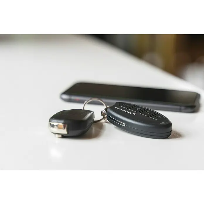Homematic IP Funkfernbedienung HmIP-KRCK - Passend für: Homematic IP Türschlossantrieb, Schwarz, 75 x 38 x 14 mm - Nahaufnahme eines Homematic-Schlüsselanhängers und eines Handys auf einem Tisch.