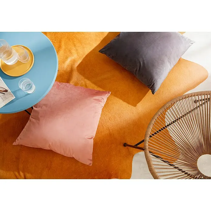Esbeco Kuhfell - Gelb, 100 % Echtfell - Orangefarbener Teppich mit Dekokissen in Rosa und Grau, ein blauer Tisch mit Gläsern und ein Stuhl mit Rohrgeflecht sorgen für Gemütlichkeit.