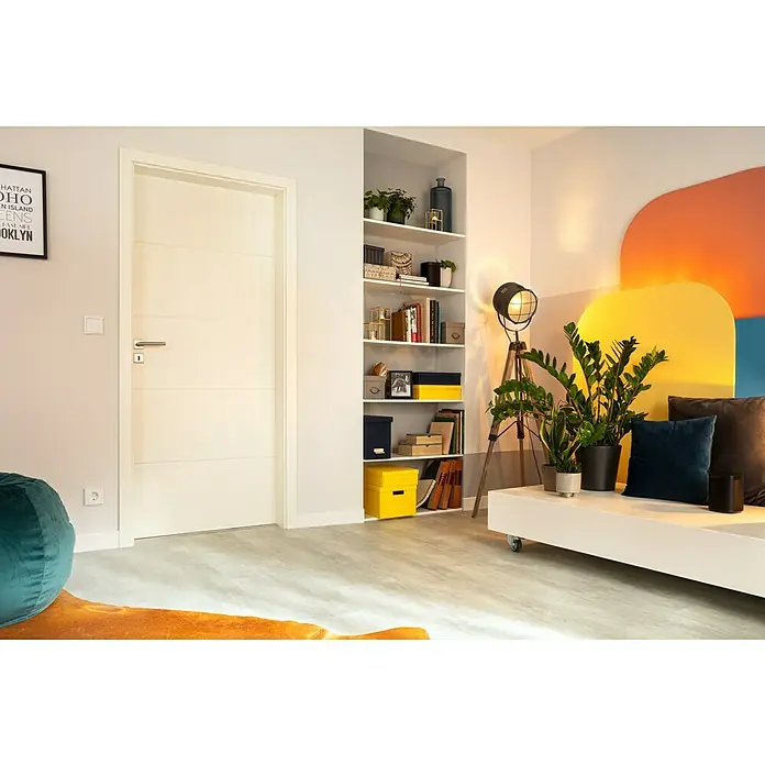 Kilsgaard Türzarge Typ 3 - 860 x 1.985 mm, Wandstärke: 290 mm, DIN Anschlag: Links, Weiß lackiert, ähnlich RAL 9010 - Elegantes Interieur mit einer weißen Tür, einem eingebauten Bücherregal mit bunten Akzenten und einem modernen Sofa mit Pflanzen und Kissen.