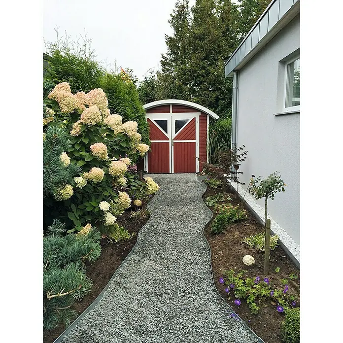 ACO Self Stabilisierungsmatte Kieswabe Eco - Polypropylen, 80 x 60 cm - Ein Gartenweg mit Kiesbelag führt zu einem roten Schuppen, umgeben von üppigen Pflanzen und Blumen. Ein idyllischer Anblick für Gartenliebhaber.