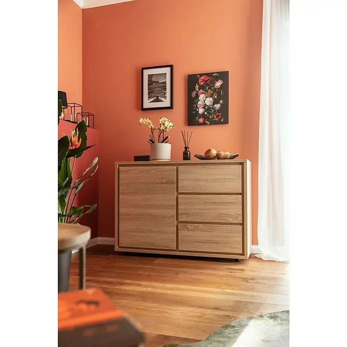 Phönix Kommode Vancouver - L x B x H: 45 x 116 x 84 cm, Eiche, Anzahl Schubladen: 3 Stk. - Ein heller Holzschrank mit Schubladen und einer Tür, dekoriert mit Blumen, Bildern und Obst, steht in einem Raum mit orangefarbenen Wänden.