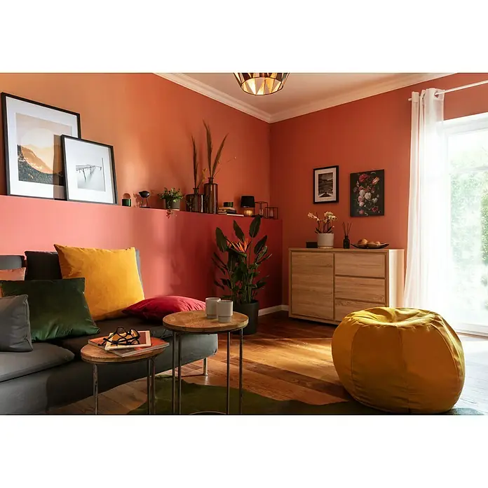 Alpina Wandfarbe Feine Farben - Befreiter Feuervogel, Matt, 2,5 l, Konservierungsmittelfrei - Wohnzimmer mit orangefarbenen Wänden, Sofa mit Kissen und Holztisch mit Brille und Tablet, wodurch ein gemütlicher und einladender Raum entsteht.