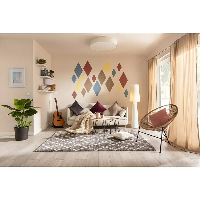 SCHÖNER WOHNEN-Farbe Wandfarbe DesignfarbeFreundliches Goldgelb, Matt, 2,5 l, Konservierungsmittelfrei - Wohnzimmer mit geometrischem Teppich in Grau, Sofa mit Kissen, Rattansessel und dekorativer Wandgestaltung.