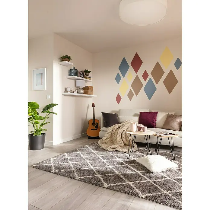 SCHÖNER WOHNEN-Farbe Wandfarbe Designfarbe - Leichtes Leinenbeige, Matt, 2,5 l, Konservierungsmittelfrei - Wohnzimmer mit grauem Teppich mit Rautenmuster, Sofa mit Kissen, Gitarre und Pflanze im Topf, der eine gemütliche Atmosphäre schafft.