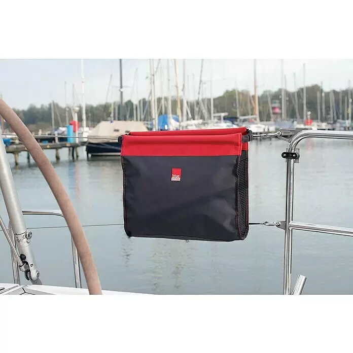 Blue Performance Relingtasche - L x B x H: 250 x 300 x 120 mm, Schwarz/Rot - Schwarze und rote Boots-Organizer-Tasche hängt an einer Edelstahlreling, mit Yachthafen und Booten im Hintergrund.