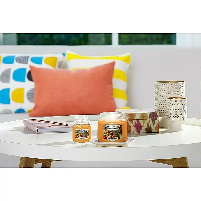 Yankee Candle Home Inspirations Duftkerze - Im Glas, Exotic Fruits, Medium - Zwei Yankee Candle Home Inspiration Exotic Fruits Kerzen, eine angezündet, auf einem weißen Couchtisch in einem Wohnzimmer mit bunten Kissen.