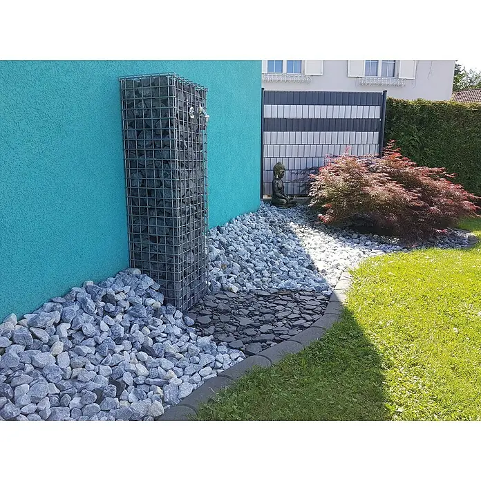 Min2C Bruchsteine - Donaublau, Körnung: 30 mm - 60 mm, 25 kg - Ein mit Steinen gefüllter Gabionen-Turm steht vor einer türkisfarbenen Wand, umgeben von dekorativen Steinen.