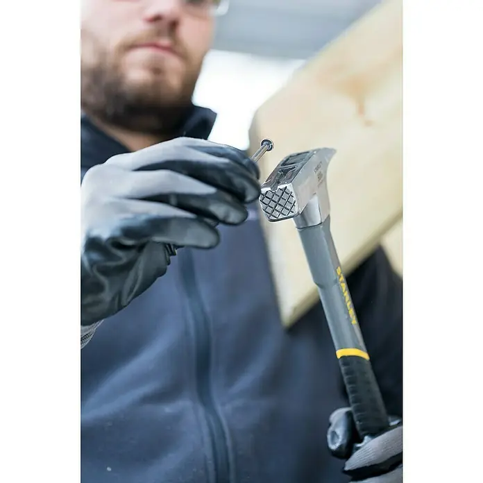 Stanley Zimmermannshammer STHT0-51312 - Gewicht Kopf: 600 g, Geraute Bahn - Nahaufnahme eines Mannes, der mit einem Stanley-Hammer und einem Nagel arbeitet und dabei Handschuhe trägt.
