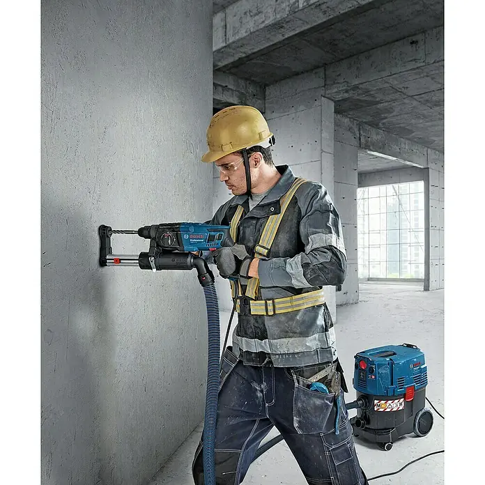 Bosch Professional Kombihammer GBH 2-21 - 720 W, 2 J, SDS-Plus-Aufnahme, Inkl. Koffer - Ein Bauarbeiter bohrt mit einer blauen Bosch Bohrhammer-Maschine ein Loch in eine Betonwand, während ein Staubsauger den Staub absaugt.