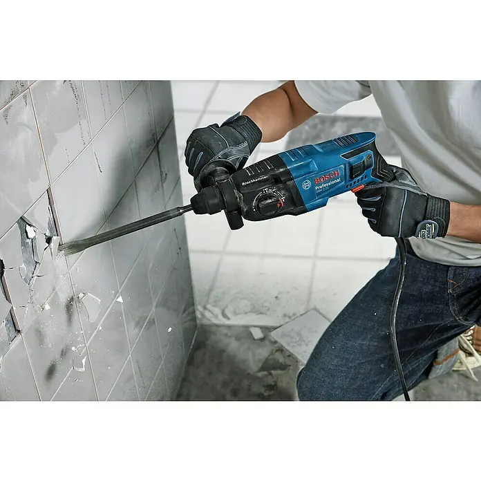 Bosch Professional Kombihammer GBH 2-21 - 720 W, 2 J, SDS-Plus-Aufnahme, Inkl. Koffer - Ein Mann entfernt mit einem Boschhammer Fliesen von einer Wand, wobei er professionelle Schutzkleidung trägt.