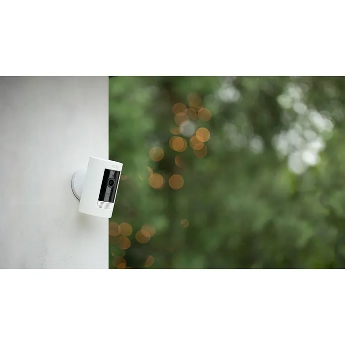 Ring Überwachungskamera außen Stick Up Cam Battery - Akkubetrieben, 1.080 Pixel (Full HD), Weiß - Eine Ring Stick Up Cam Battery, eine kabellose Sicherheitskamera in Weiß, ist an einer weißen Wand mit grünem Hintergrund befestigt.