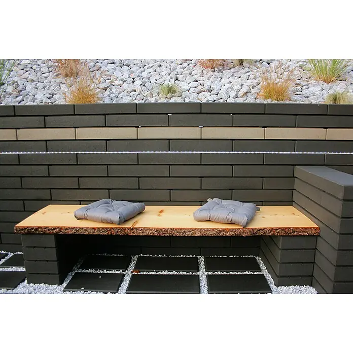Christoph Mauerstein C-Line - Anthrazit, 50 x 25 x 10 cm, Beton - Gartenbank mit zwei grauen Kissen auf einer Holzoberfläche vor einer Steinmauer, die mit einer LED-Leiste beleuchtet wird.