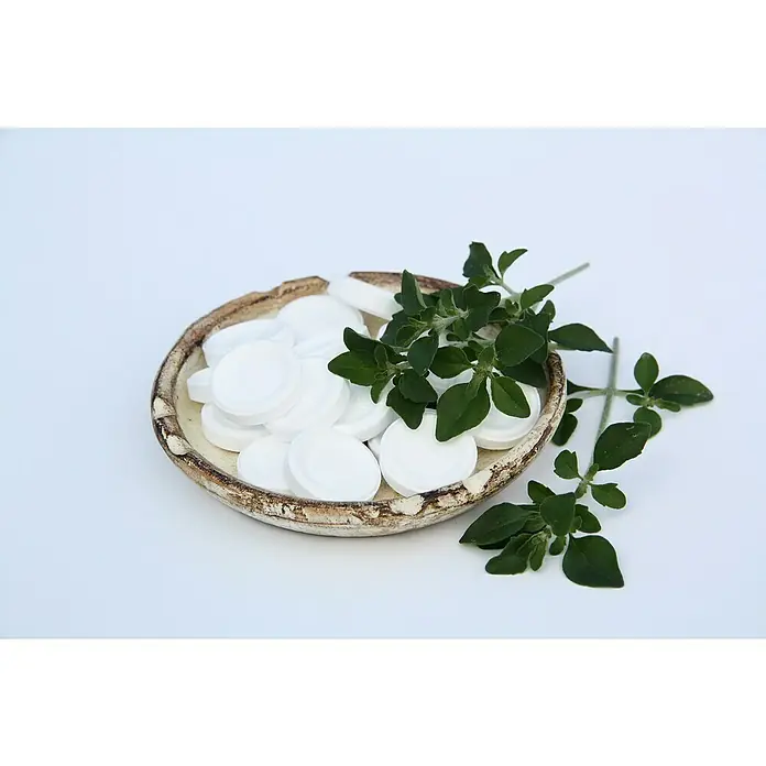 Blu Bio Minzbusch - Prostanthera rotundifolia - Nahaufnahme eines rustikalen Tellers mit weißen Tabletten und frischen grünen Kräutern auf einem sauberen, hellen Hintergrund.