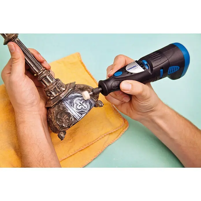 Dremel Fieltro de pulido 414 - Diámetro: 13 mm, 6 ud. - Primer plano de una persona puliendo un objeto metálico ornamentado con una herramienta Dremel 7.2V, con un paño amarillo debajo.