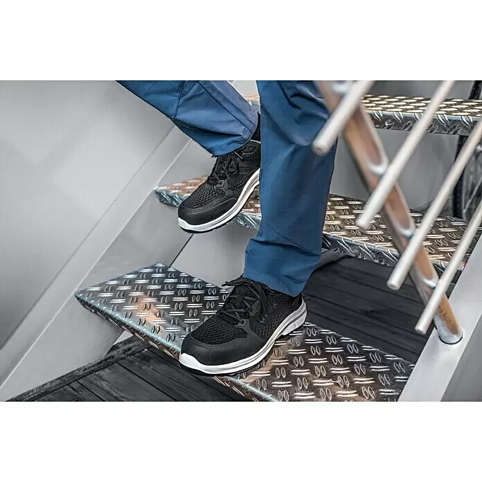 Uvex Sicherheitshalbschuh 1 Sport - 45, Schutzklasse nach EN ISO 20345: S1 - Person mit schwarzen und weißen Uvex-Sneakern und blauen Hosen, die Metalltreppen hinuntergeht.