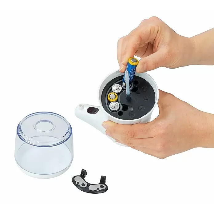 Wenko Desinfektionsmittelspender Diala - Weiß, L x B x H: 19 x 12 x 23 cm - Hände setzen Batterie in weiß-schwarzen Automatikspender ein. Klarer Behälter und Batteriedeckel liegen daneben.