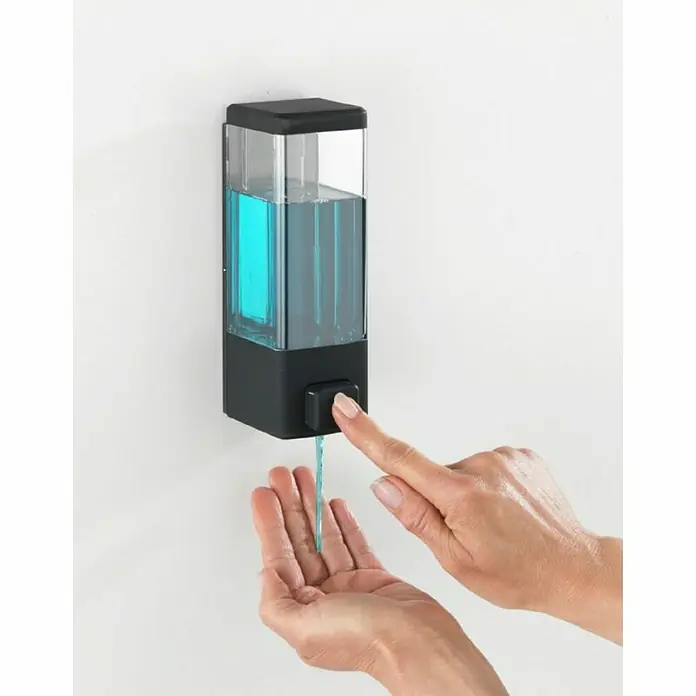 Wenko Dispensador de jabón o gel desinfectante Istres - Plástico, Negro, Capacidad del depósito: 320 ml - Esta imagen muestra un elegante dispensador de jabón negro y transparente montado en una pared, con una mano dispensando el jabón azul.