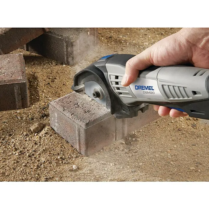 Dremel Mauerwerk-Trennscheibe DSM 520 - 77 mm, Geeignet für: Mauerwerk, Schnitttiefe: 20 mm, 2 Stk. - Eine Dremel DSM20 Kompaktsäge schneidet durch einen Pflasterstein, wobei Staub aufwirbelt. Das Gerät ist grau mit schwarzen und blauen Akzenten.