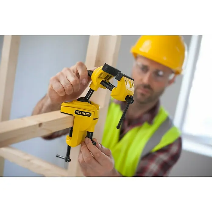 Stanley Schraubstock MaxSteel - Backenbreite: 70 mm - Der gelbe Stanley Schraubstock wird von einem Bauarbeiter mit gelbem Helm und Schutzbrille an einem Holzstück befestigt.