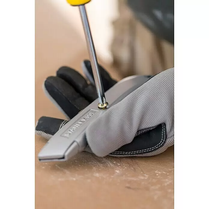 Stanley Schiebemesser 99E - Trapezklinge, Schiebbare Klinge - Eine behandschuhte Hand hält ein silbernes Teppichmesser, das mit einem gelbgriffigen Schraubendreher aufgeschraubt wird. Das Messer zeigt „STANLEY 99E“.