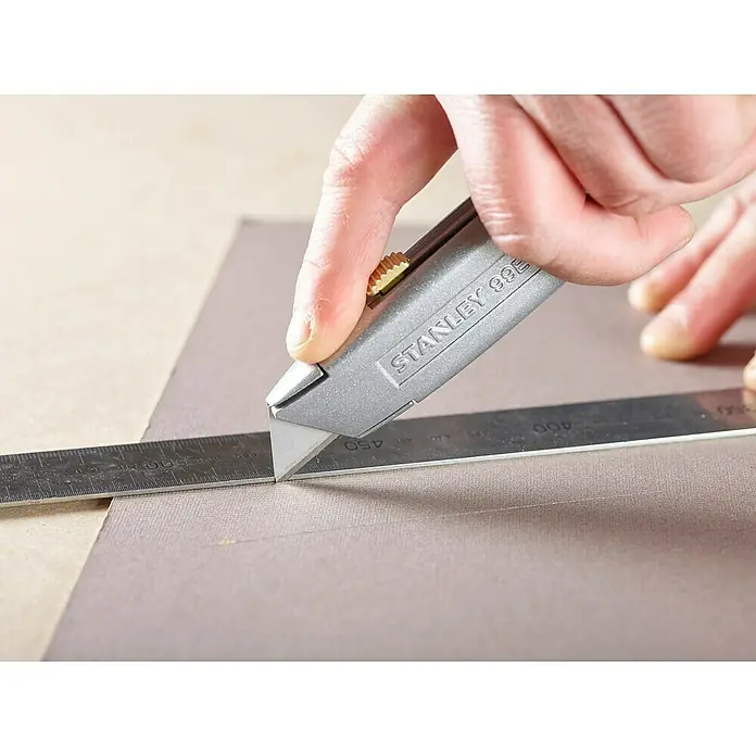 Stanley Schiebemesser 99E - Trapezklinge, Schiebbare Klinge - Nahaufnahme einer Person, die mit einem Stanley 99E Cuttermesser entlang eines Metalllineals auf einer grauen Oberfläche schneidet.