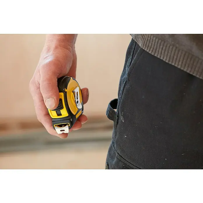Stanley Kapselbandmaß Tylon - 8 m - Ein Handwerker hält ein gelb-schwarzes Maßband in der Hand, während er Arbeitskleidung trägt, die auf eine Baustelle oder ein Heimwerkerprojekt hindeutet.