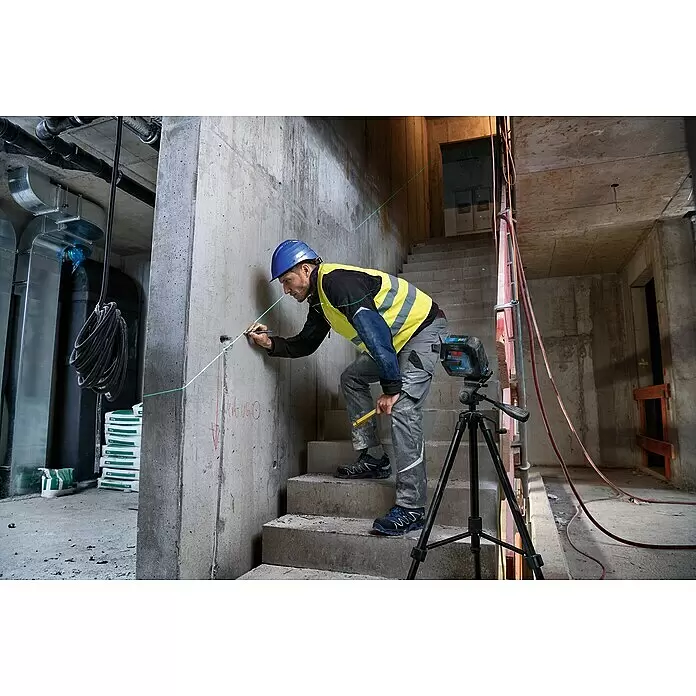 Bosch Professional Linienlaser GLL 2-15 G - Max. Arbeitsbereich: 15 m, 5 -tlg. - Bosch Laser-Nivelliergerät auf Stativ, projiziert grüne Linien, von einem Arbeiter zum Markieren einer Betonwand auf einer Baustelle verwendet.