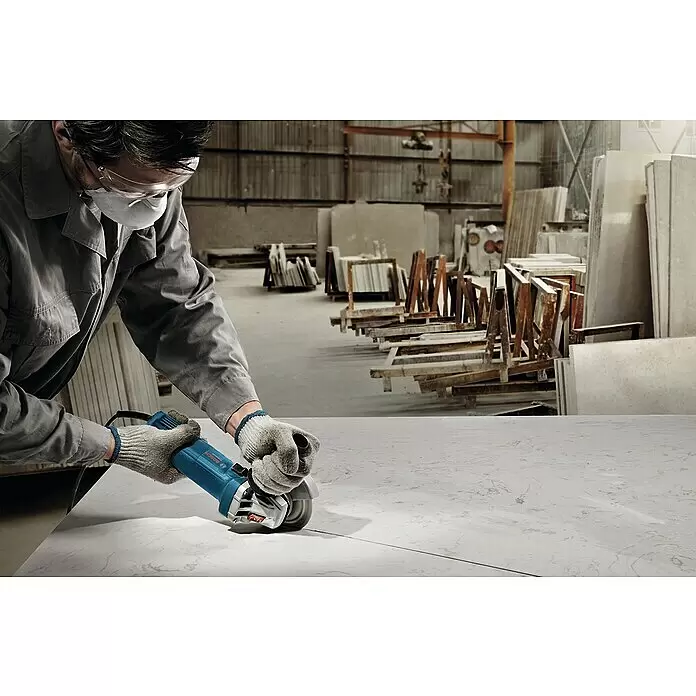Bosch Professional Elektro-Winkelschleifer GWS 900 - 900 W, Durchmesser Scheibe: 125 mm, Leerlaufdrehzahl: 11.000 U/min - Person in Schutzkleidung schneidet mit blauem Winkelschleifer eine weiße Platte in einer Werkstatt.