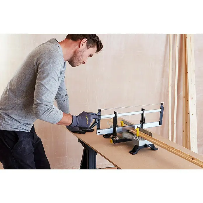 Stanley Gehrungslade 1-20-800 mit Säge - L x B x H: 560 x 143 x 95 mm - Ein Mann schneidet Holz mit einer Stanley-Säge, wobei er Handschuhe trägt. Die Säge steht auf einem Holztisch.