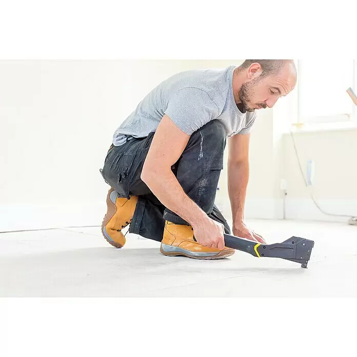 Stanley FatMax Martillo grapador FMHT81394-9 - Se puede recargar - Un hombre usa un estirador de alfombras negro y amarillo, arrodillado en un suelo claro para estirar alfombras o pisos.