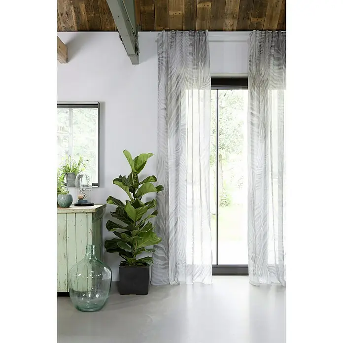 Rasch Home Schlaufenbandschal Tahiti - 140 x 255 cm, 100 % Polyester, Grau/Grün - Wohnraum mit hellem Vorhang mit Blattmuster vor großem Fenster, grüner Pflanze im Topf und Holzbalken an der Decke.