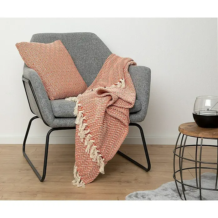 Decke Zigzag - Rostrot, 170 x 130 cm, 100 % Baumwolle - Nahaufnahme eines grauen Sessels mit einem orangefarbenen Kissen und einer passenden Decke mit Fransen.