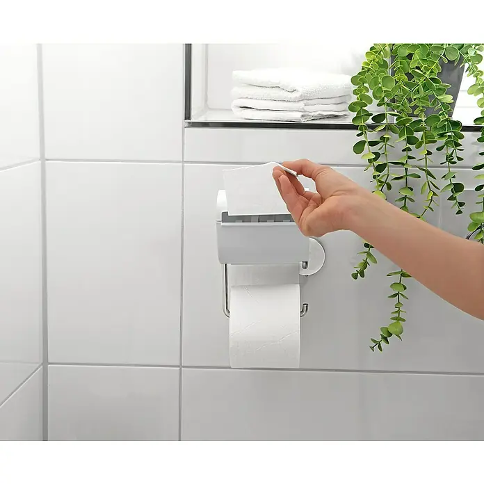 Toilettenpapierbefeuchter bideo - Weiß/Chrom, Glänzend - Eine Hand zieht ein Blatt Papier aus einem weißen Papierhalter im Badezimmer mit weißen Fliesen und einer Pflanze im Hintergrund.