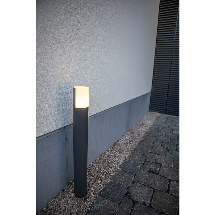 Lutec Sobremuro exterior Cyra - 8 W, Color del marco: Gris oscuro, Blanco cálido, 78 x 78 x 650 mm, Con bombillas - Una luz exterior moderna en forma de pilar ilumina un camino pavimentado frente a una pared clara. La luz emite un brillo cálido.