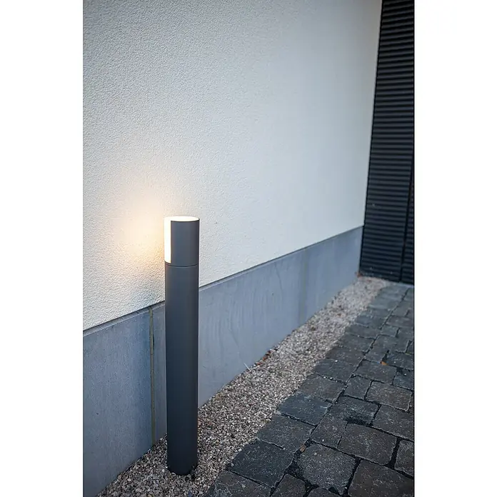 Lutec Sobremuro exterior Cyra - 8 W, Color del marco: Gris oscuro, Blanco cálido, 78 x 78 x 650 mm, Con bombillas - Una luz de camino metálica gris con aberturas rectangulares e iluminación integrada se encuentra junto a una pared de piedra gris.