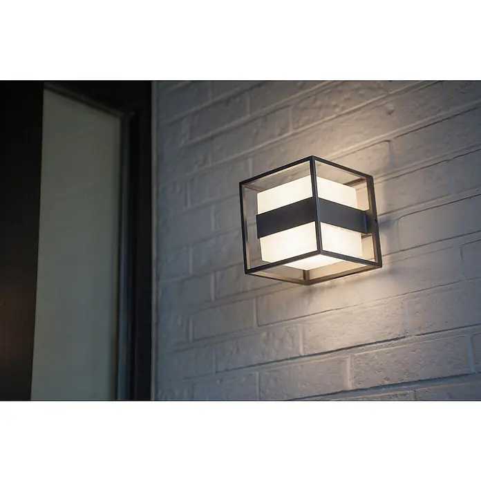 Lutec Aplique exterior LED Cruz - 13 W, 140 x 140 x 148 mm, Gris oscuro, IP54 - Un moderno aplique exterior en forma de cubo hecho de metal y vidrio en una pared de ladrillo blanco, que irradia una luz cálida y acogedora.
