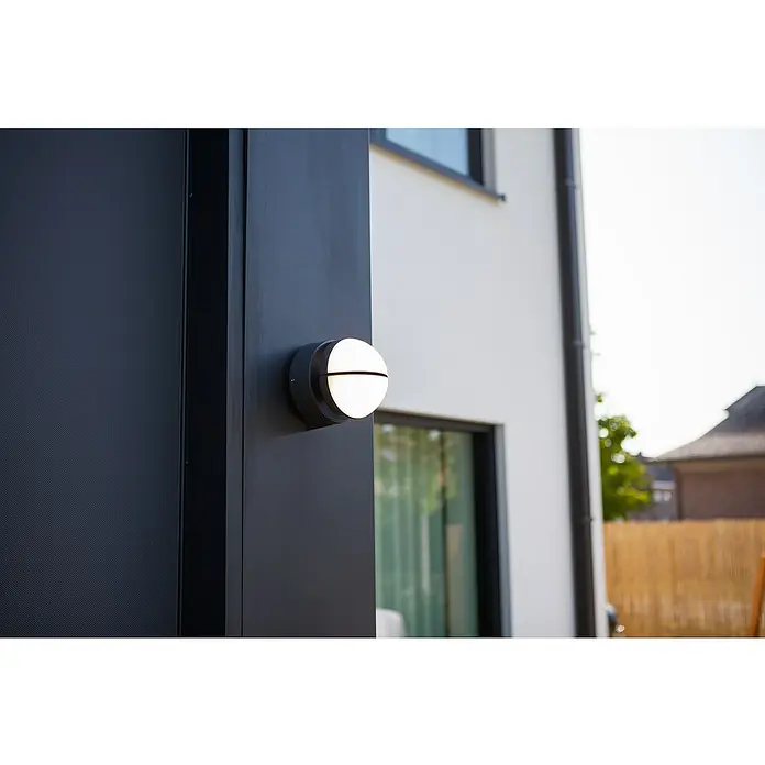 Lutec Aplique exterior LED Eklips - 13 W, 161 x 161 x 146 mm, Gris oscuro, IP54 - Un primer plano muestra una luminaria exterior redonda con una carcasa negra y un difusor iluminado dividido horizontalmente, montado en una columna oscura.