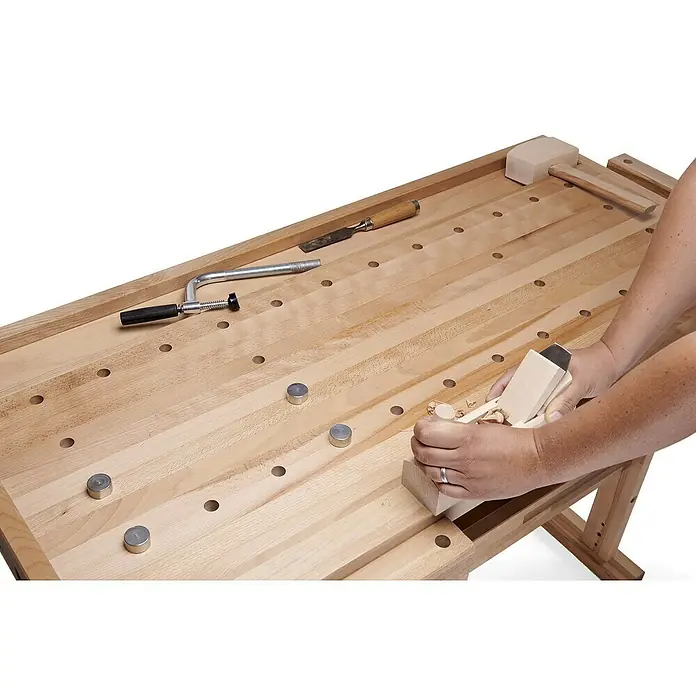 Ramia Hobelbank Diamant 1800 - L x B: 180 x 76 cm, Arbeitshöhe: 85 cm, Holz - Ein Holzarbeiter benutzt eine Hobelbank mit verschiedenen Werkzeugen, um Holz zu bearbeiten. Die Werkbank ist hell und weist Löcher für die Werkzeuge auf.